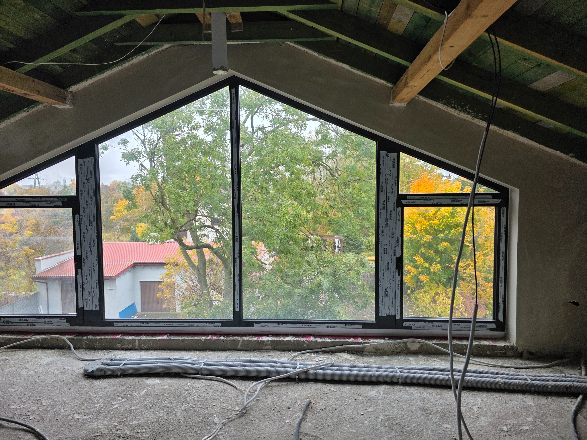 Okno dachowe z czarnymi ramami w surowym wnętrzu poddasza. Widok na drzewa w jesiennych barwach. Na podłodze rury instalacyjne.