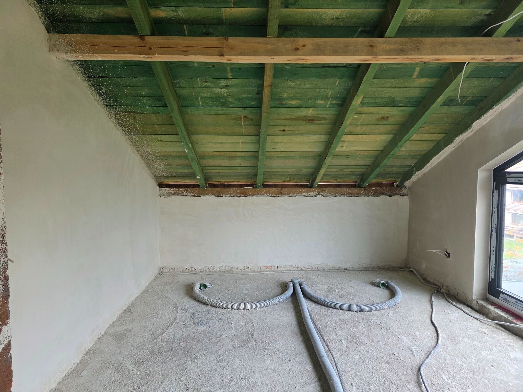 Pokój w trakcie remontu: surowe ściany po szpachlowaniu, zielone drewniane belki stropowe, szare rury wentylacyjne na podłodze. Widok na okno.