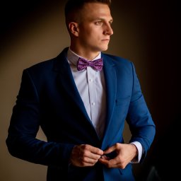 Dorian Kapiszewski Fotografia - Młody mężczyzna w niebieskim garniturze i wzorzystej muszce, zapinający marynarkę, patrzy w bok na ciemnym tle.