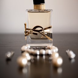 Dorian Kapiszewski Fotografia - Perfumy w kwadratowym flakonie z czarnym korkiem i złotym logo YSL, leżące na ciemnym blacie z rozproszonym naszyjnikiem z pereł w tle.