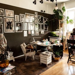 Dorian Kapiszewski Fotografia - Wnętrze studia z ciemną ścianą ozdobioną czarno-białymi fotografiami, gdzie wizażysta poprawia makijaż klientki siedzącej na krześle przy oknie, a obok stoi stół z kosmetykami i lampą.