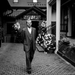 Dorian Kapiszewski Fotografia - Czarno-białe zdjęcie mężczyzny w garniturze spacerującego po brukowanej ścieżce przed budynkiem z napisem 'Stary Lwów' i kwiatami w donicach.