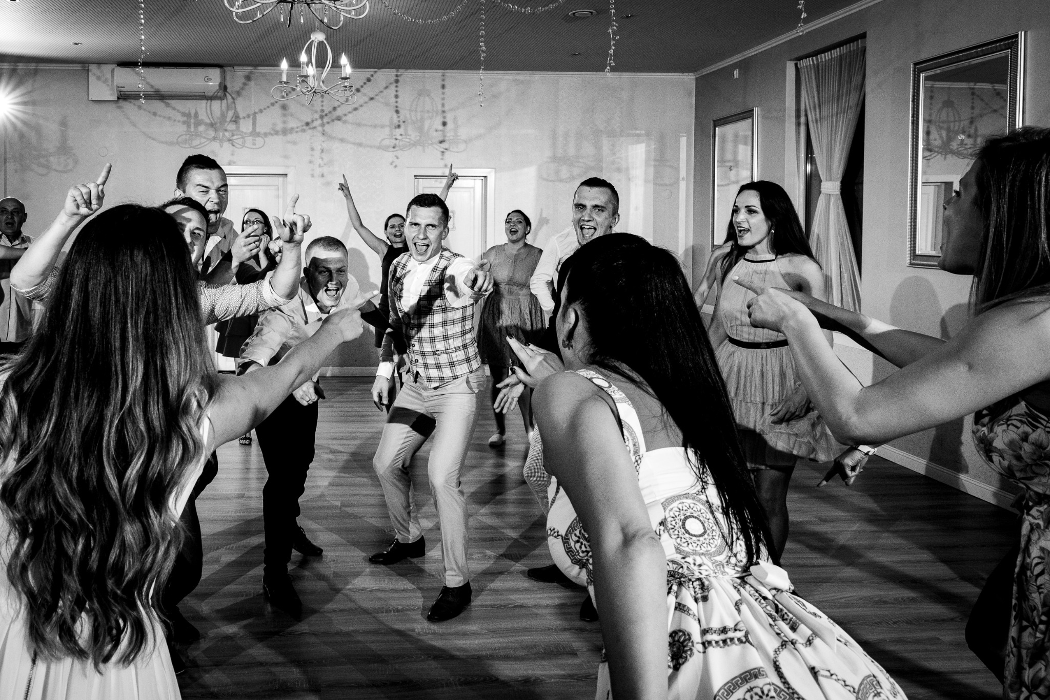 Czarno-białe zdjęcie z imprezy: grupa rozbawionych osób tańczy i wskazuje palcami, oświetlona żyrandolem, z parkietem pod nogami.