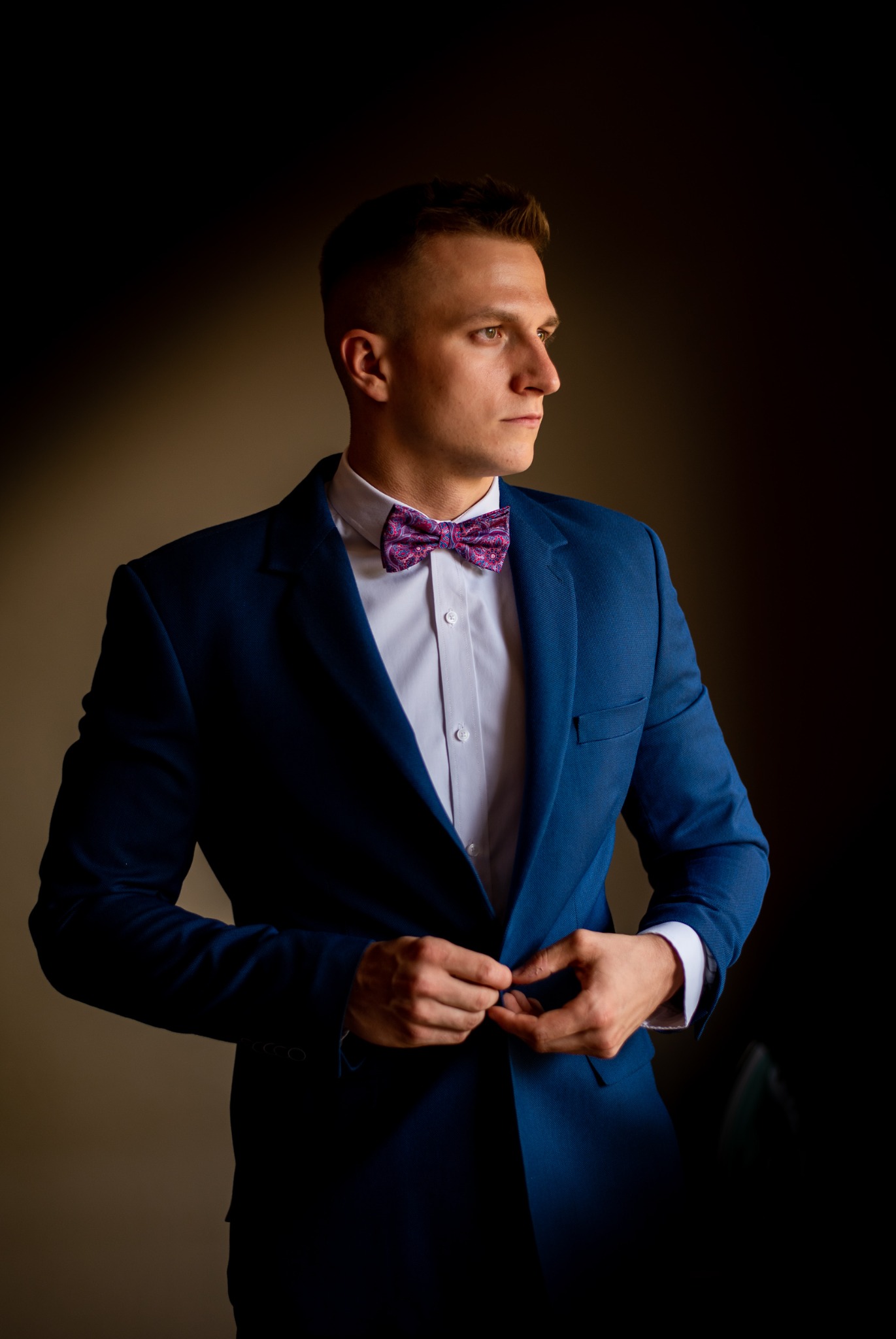 Młody mężczyzna w niebieskim garniturze i wzorzystej muszce, zapinający marynarkę, patrzy w bok na ciemnym tle.