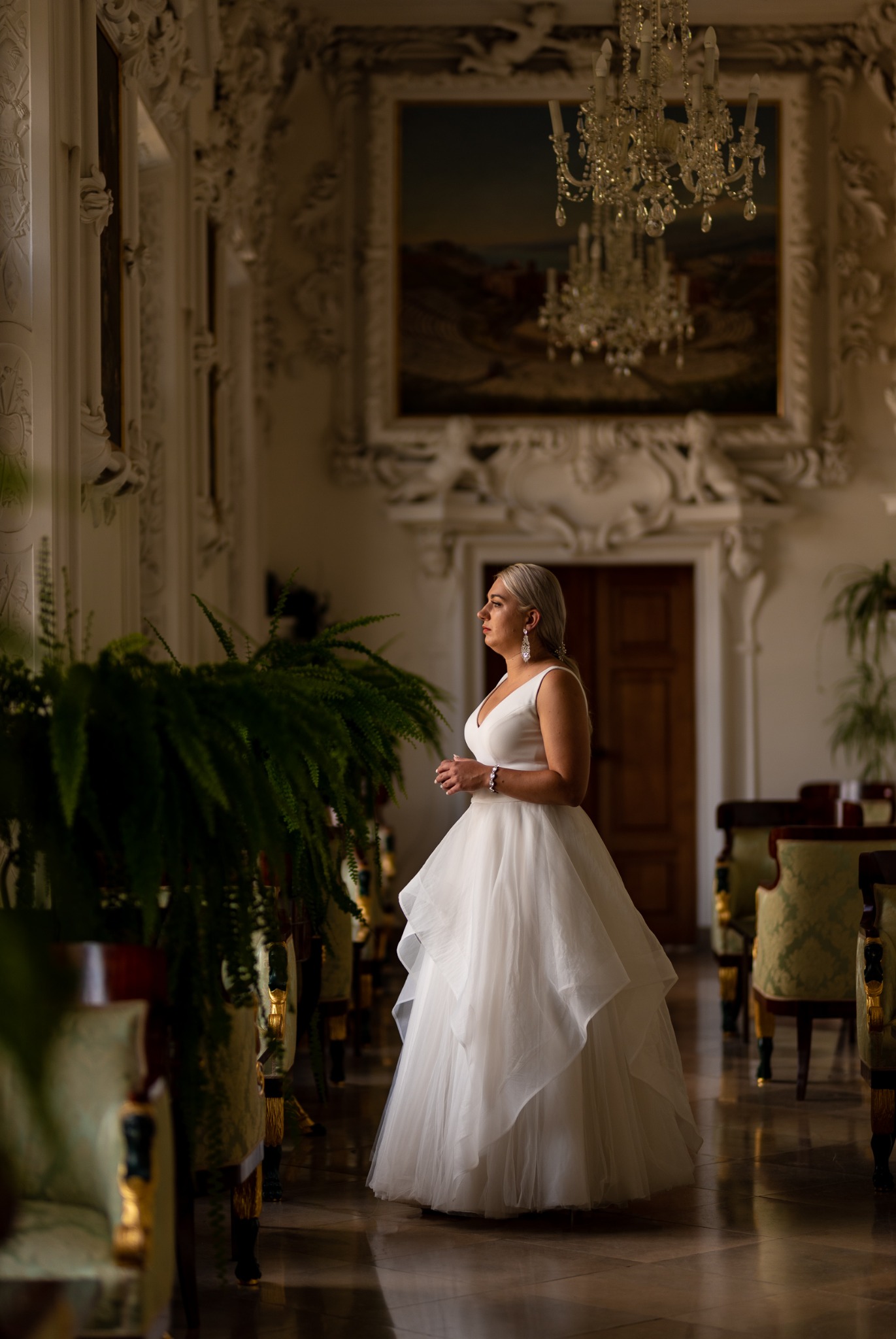 Panna młoda w białej sukni, spoglądająca przez okno w bogato zdobionym wnętrzu z żyrandolem i obrazem w tle.
