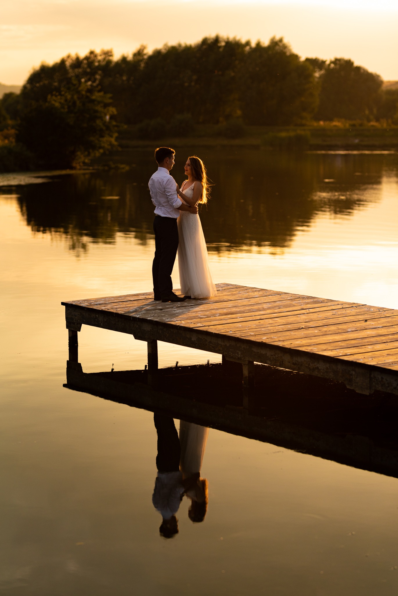 Para młoda w objęciach na drewnianym pomoście nad jeziorem o zachodzie słońca, z odbiciem w wodzie.