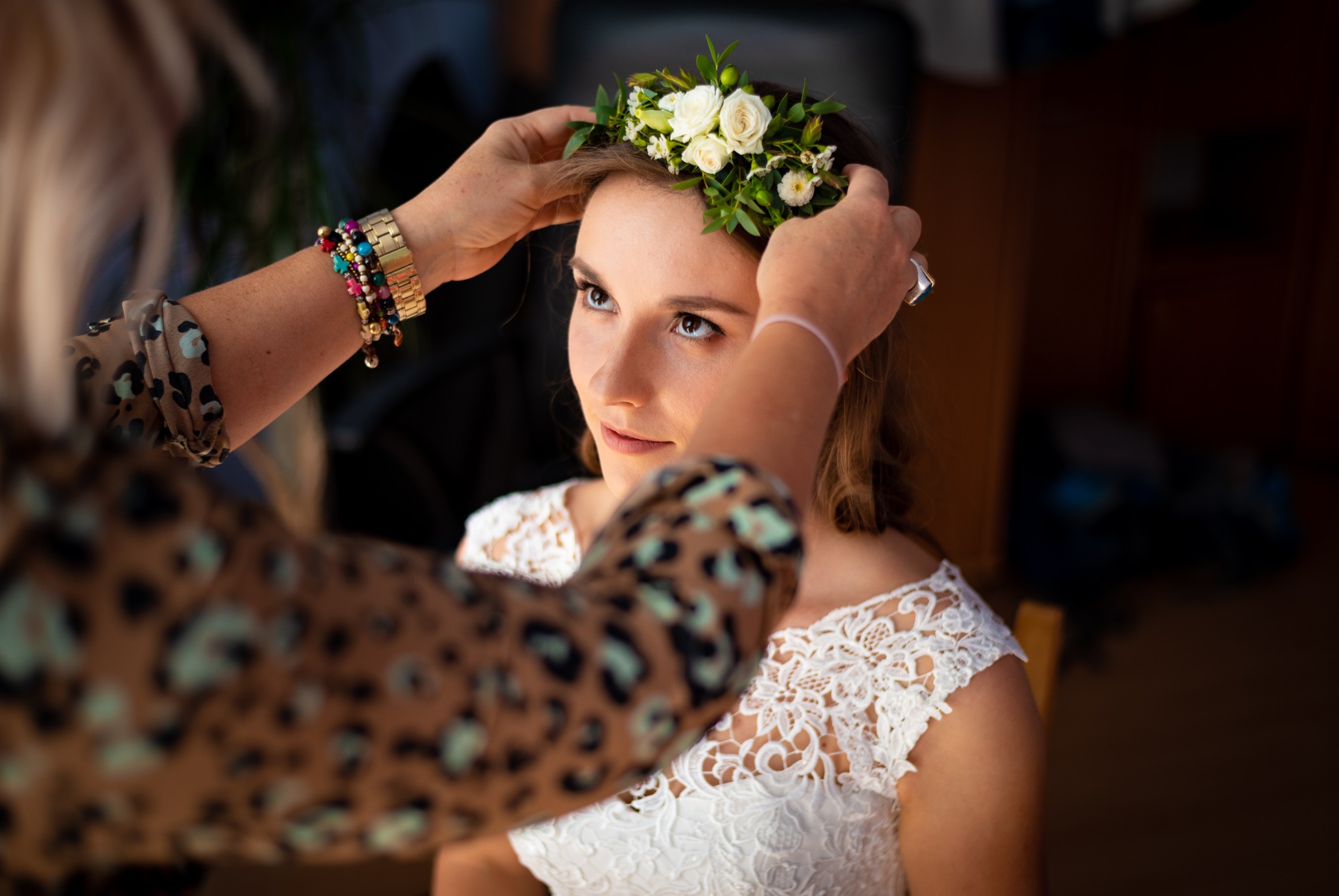 Panna młoda w białej koronkowej sukni, podczas zakładania wianka z białych róż i zielonych listków, widok z bliska na twarz i dłonie z biżuterią.