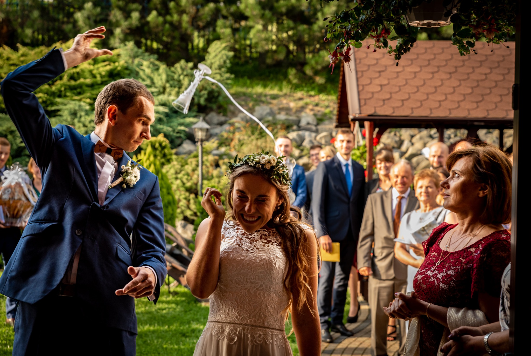 Panna młoda w koronie z kwiatów śmieje się, gdy pan młody rzuca za nią kieliszki do wina, w tle goście weselni i zielona sceneria.