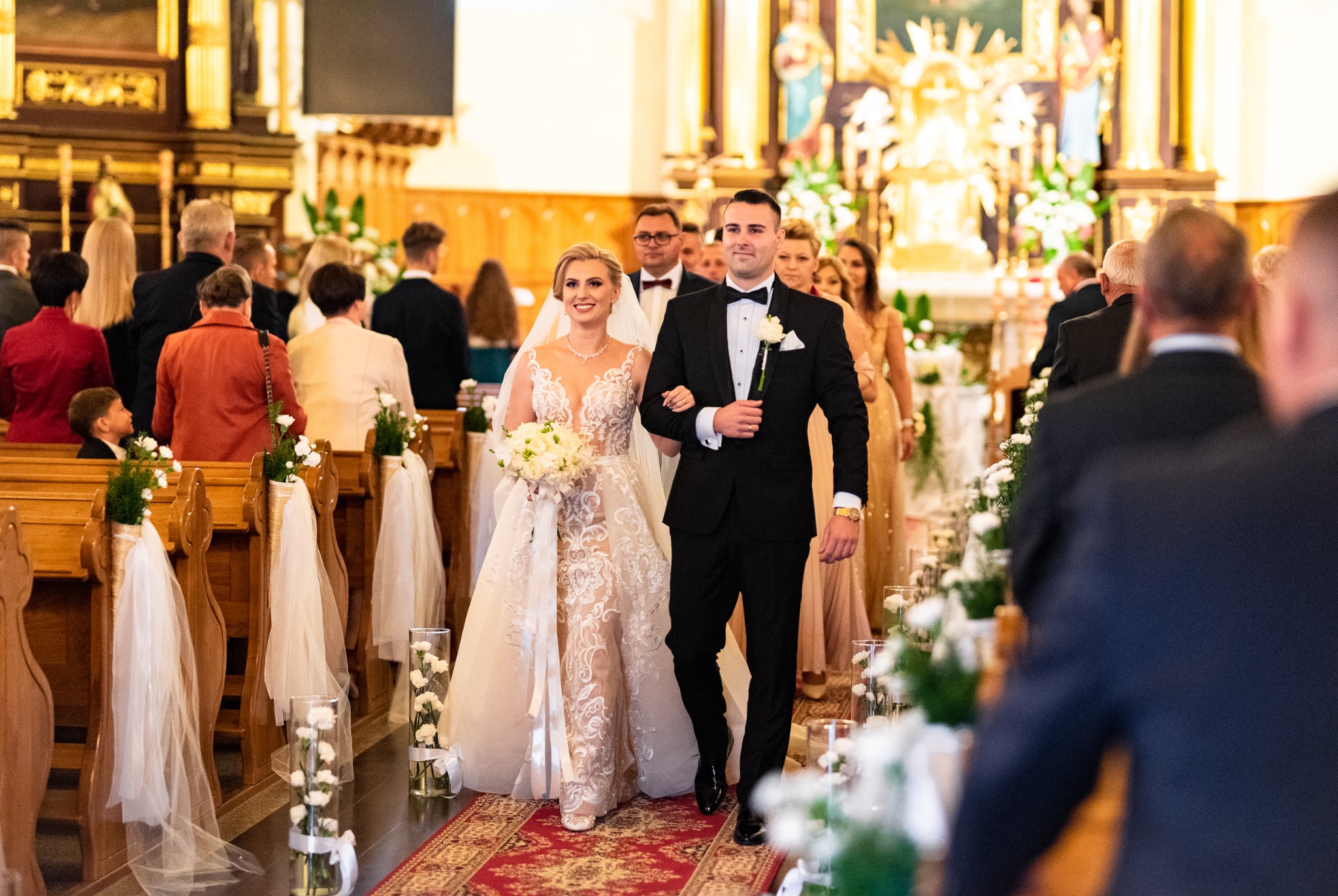 Para młoda w kościele idzie do ołtarza, panna młoda w koronkowej sukni trzyma bukiet białych kwiatów, pan młody w czarnym smokingu z butonierką, dekoracje kwiatowe wzdłuż ławek.
