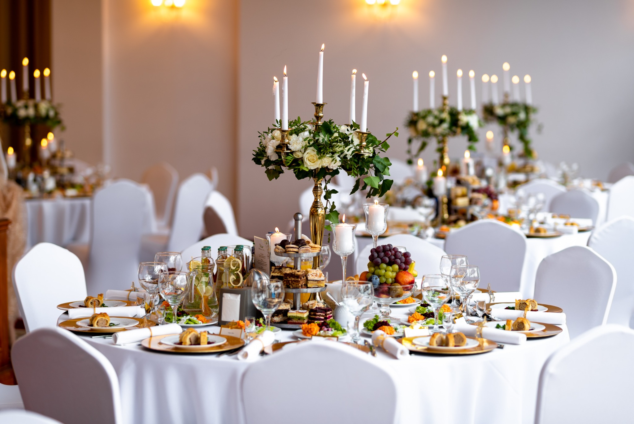 Uroczysty stół weselny z białym obrusem, złotymi talerzami, świecami w złotych świecznikach z kwiatami, szklankami, deserami i owocami, nakryty dla wielu gości.