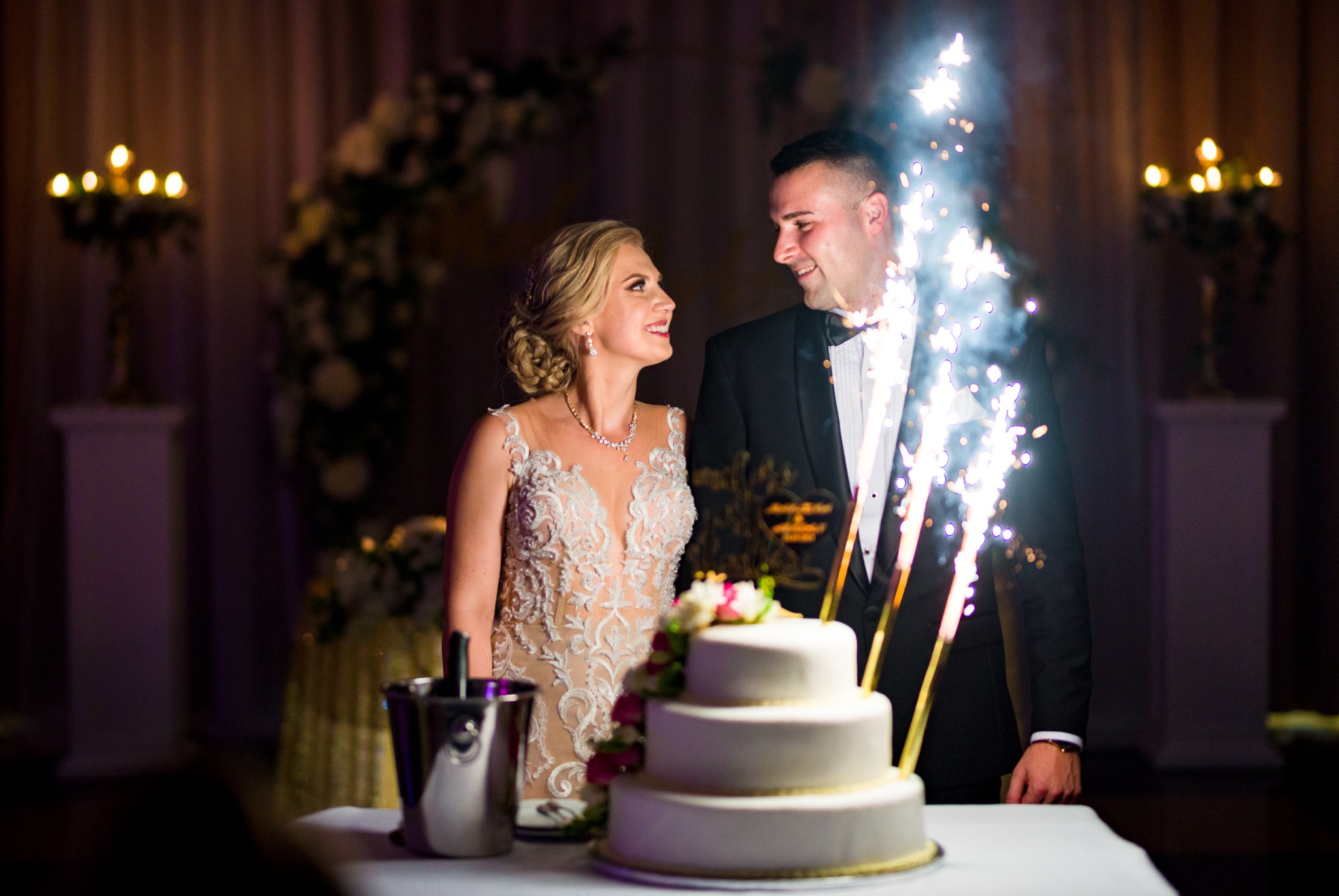 Uśmiechnięta para młoda spogląda na siebie przy trzy-piętrowym torcie weselnym, z fontannami iskier po bokach, w tle dekoracje kwiatowe i świeczniki.