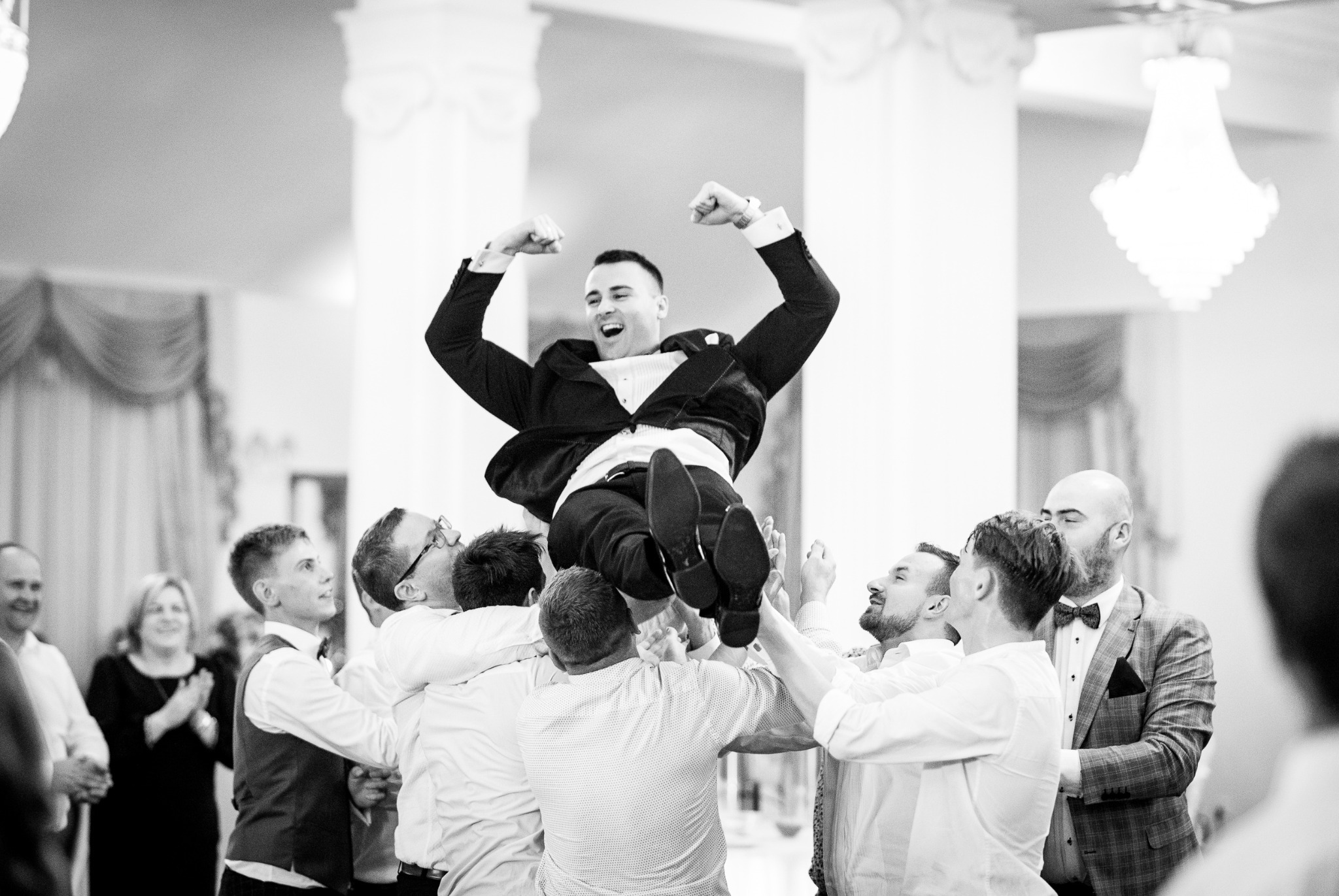 Czarno-białe zdjęcie mężczyzny w garniturze podrzucanego do góry przez grupę osób podczas uroczystości, z widocznymi kolumnami i żyrandolem w tle.