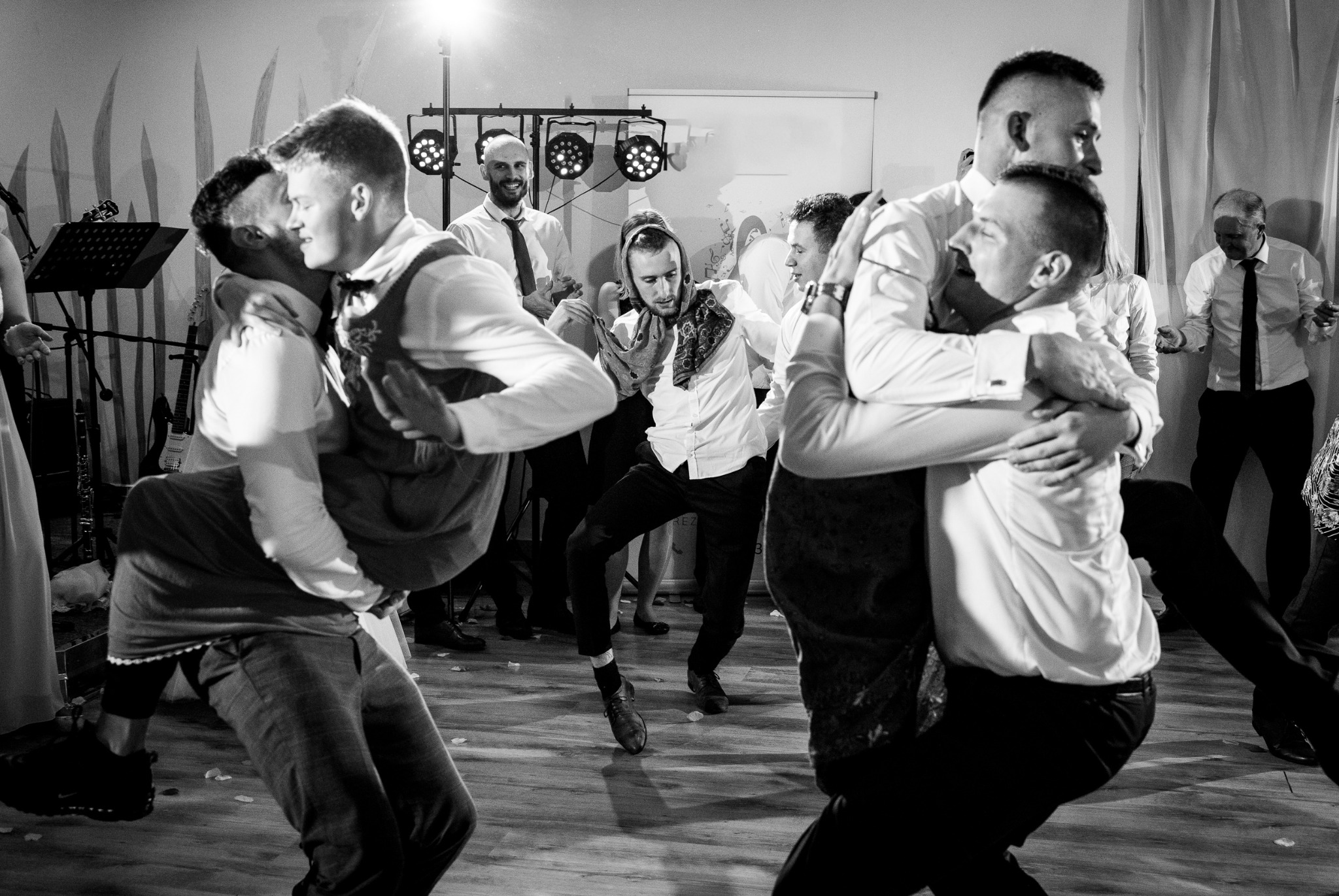 Czarno-białe zdjęcie z wesela: goście tańczą, dwóch mężczyzn podnosi innych w górę, w tle zespół muzyczny i oświetlenie sceniczne.