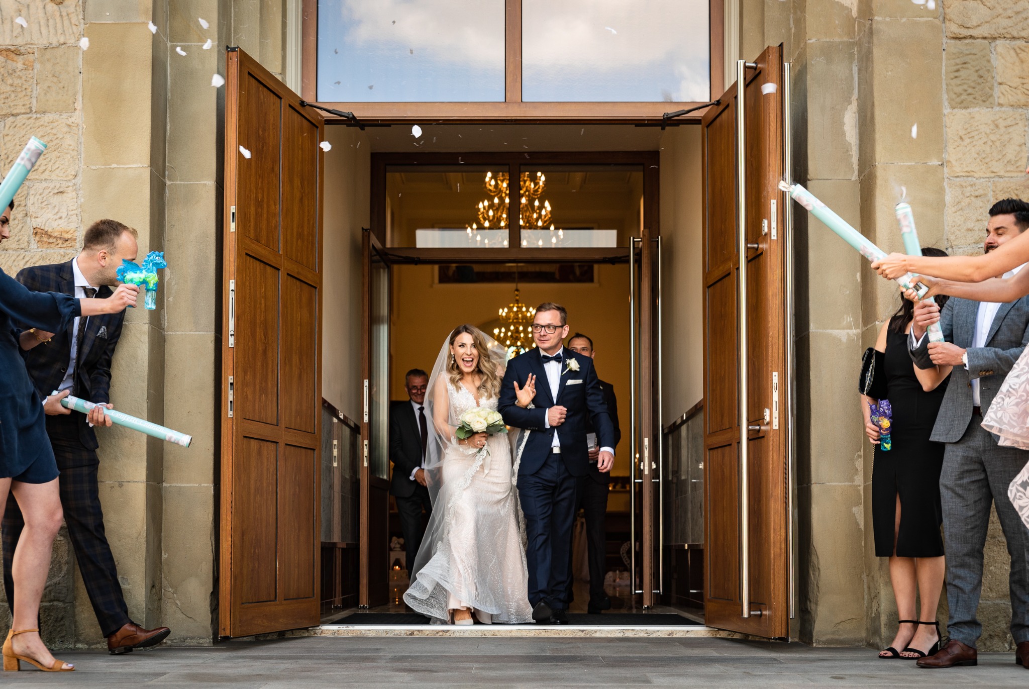 Uśmiechnięta para młoda wychodzi z kościoła, obsypywana konfetti, goście trzymają tuby i pistolety na bańki mydlane.