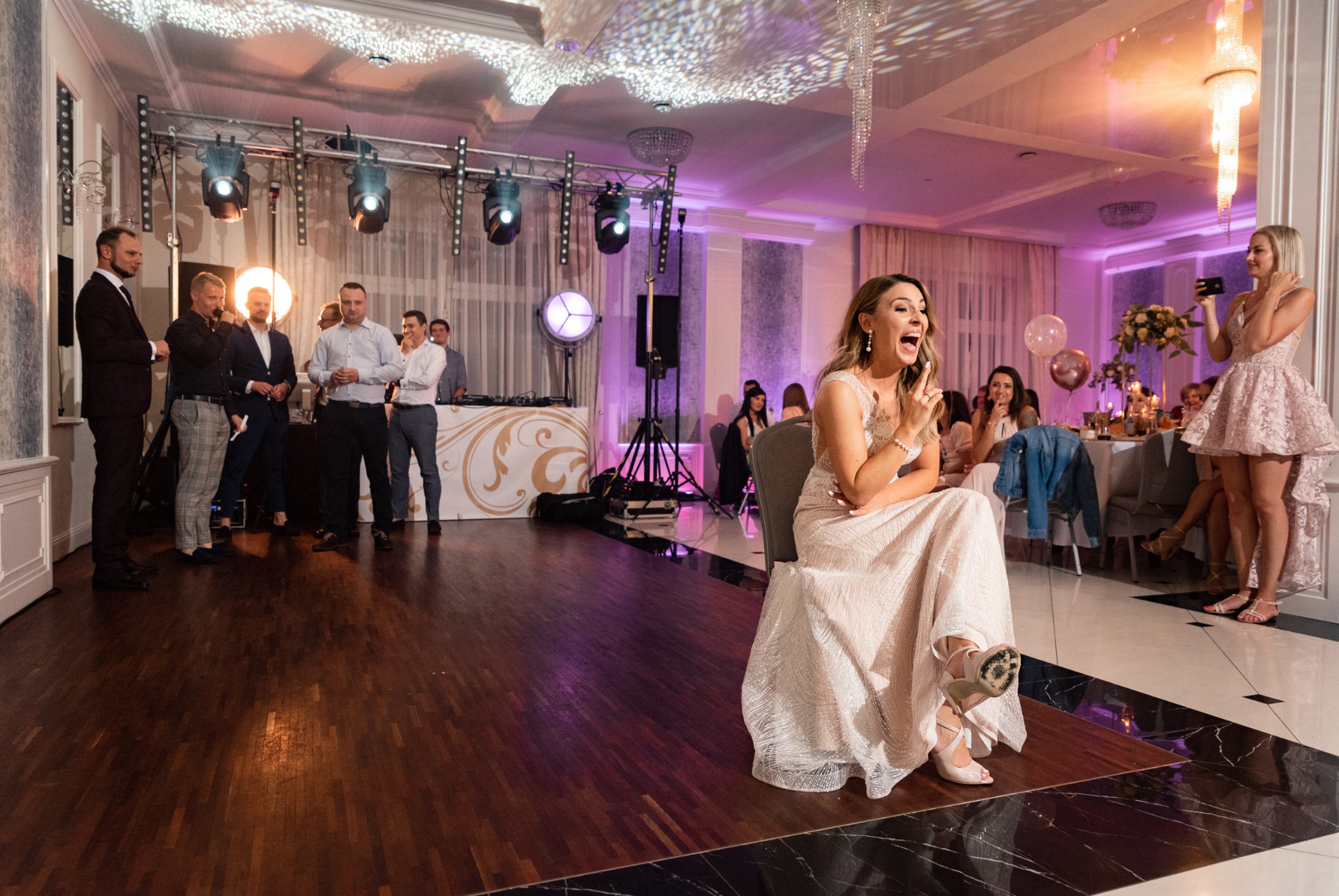 Panna młoda w sukni ślubnej siedzi na krześle, z szeroko otwartymi ustami i uniesioną ręką, w tle orkiestra i goście weselni.