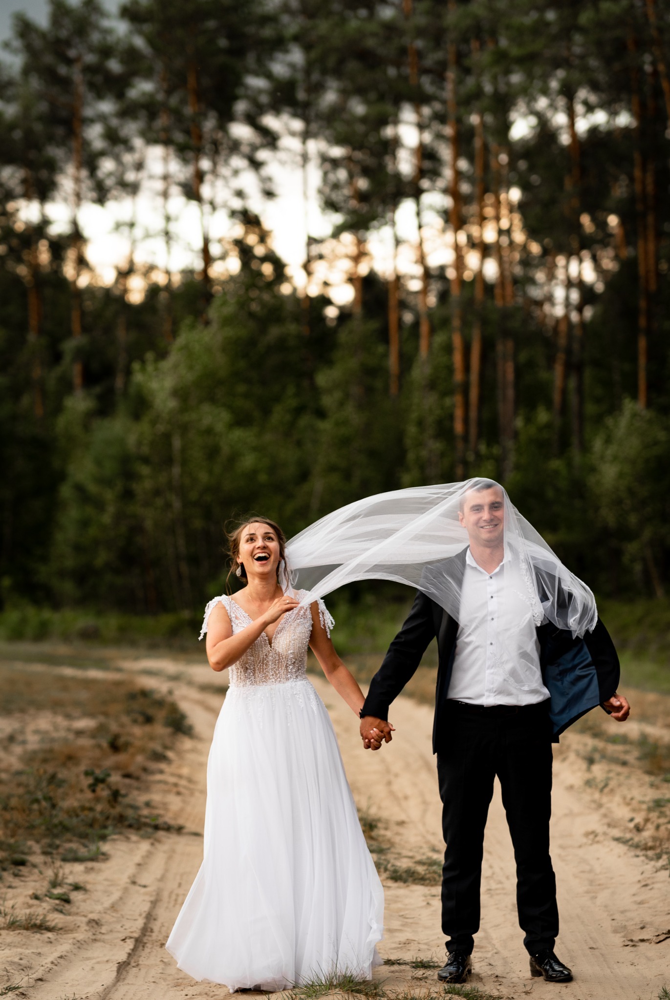 Uśmiechnięta para młoda trzymająca się za ręce na piaszczystej drodze, panna młoda w koronkowej sukni, pan młody z welonem na głowie, las w tle.