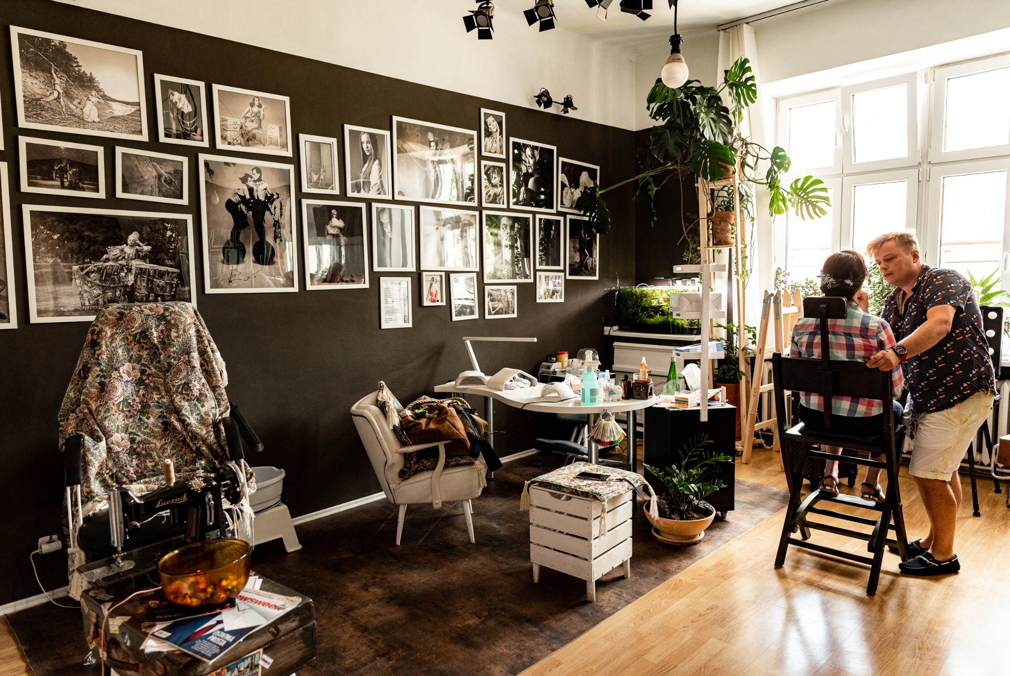 Wnętrze studia z ciemną ścianą ozdobioną czarno-białymi fotografiami, gdzie wizażysta poprawia makijaż klientki siedzącej na krześle przy oknie, a obok stoi stół z kosmetykami i lampą.