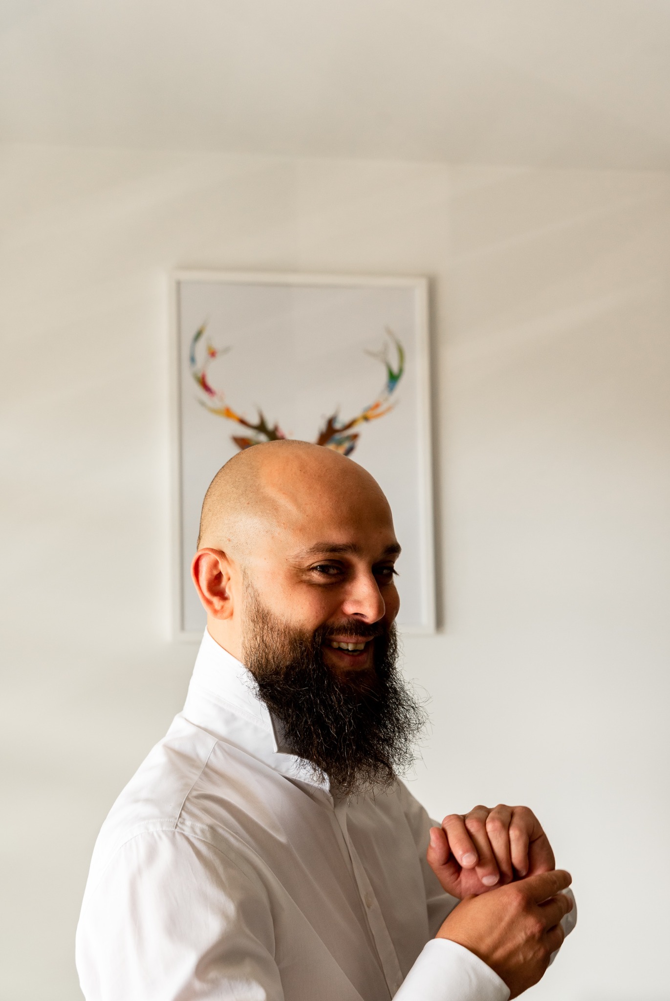 Uśmiechnięty mężczyzna z brodą w białej koszuli zapina mankiet na tle białej ściany z obrazem przedstawiającym kolorowe poroże jelenia.
