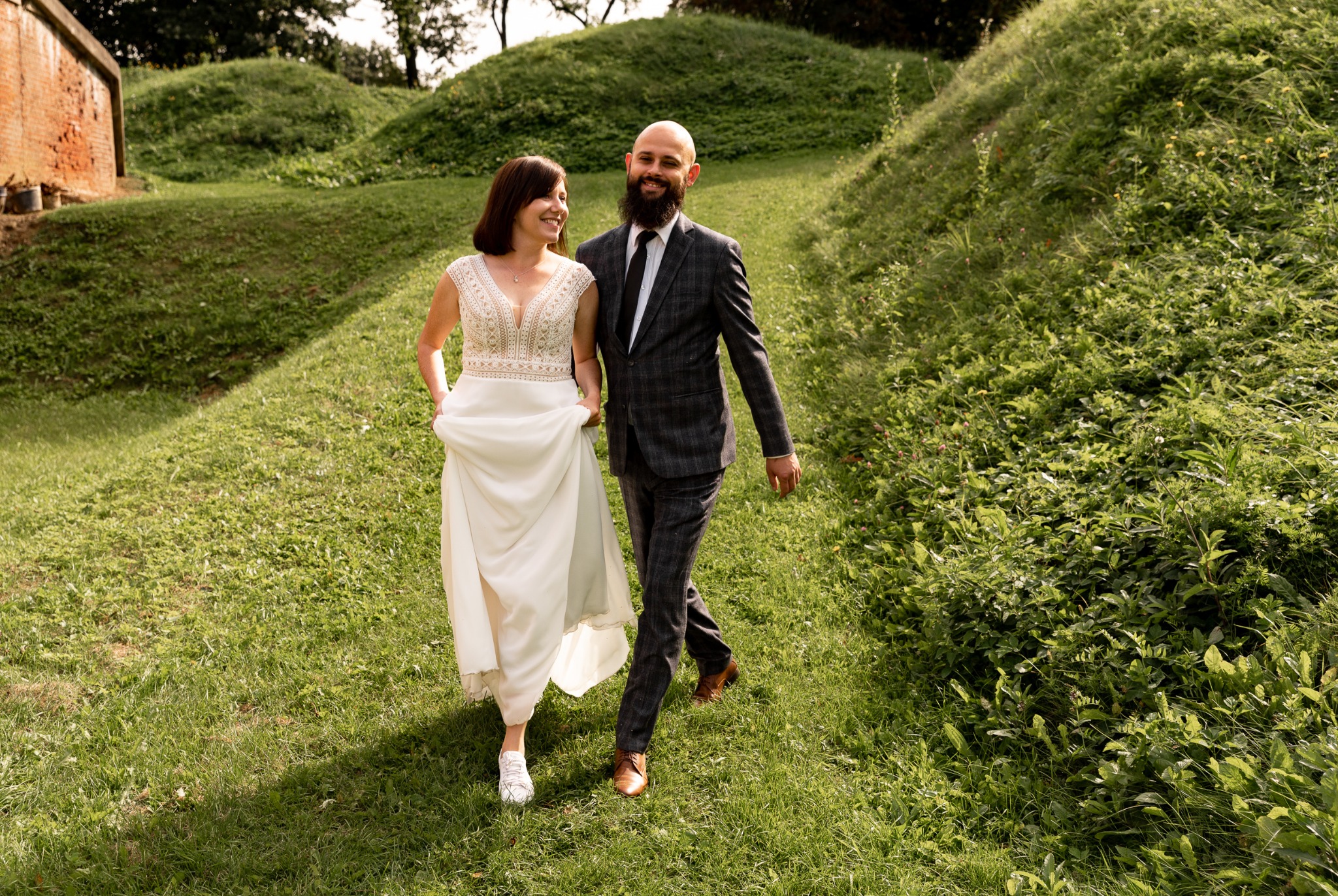 Para młoda spaceruje po trawiastym terenie: panna młoda w białej sukni z koronkową górą i sportowych butach, pan młody w szarym garniturze w kratę i brązowych butach.