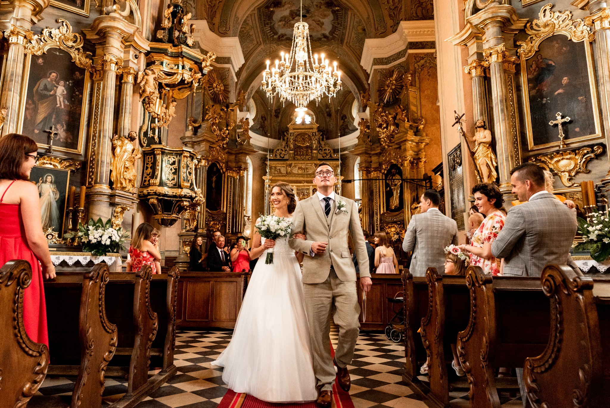 Para młoda w beżowych strojach idzie przez kościół z czarno-białą szachownicą na podłodze, otoczona gośćmi weselnymi i bogato zdobionym wnętrzem z obrazami i żyrandolem.