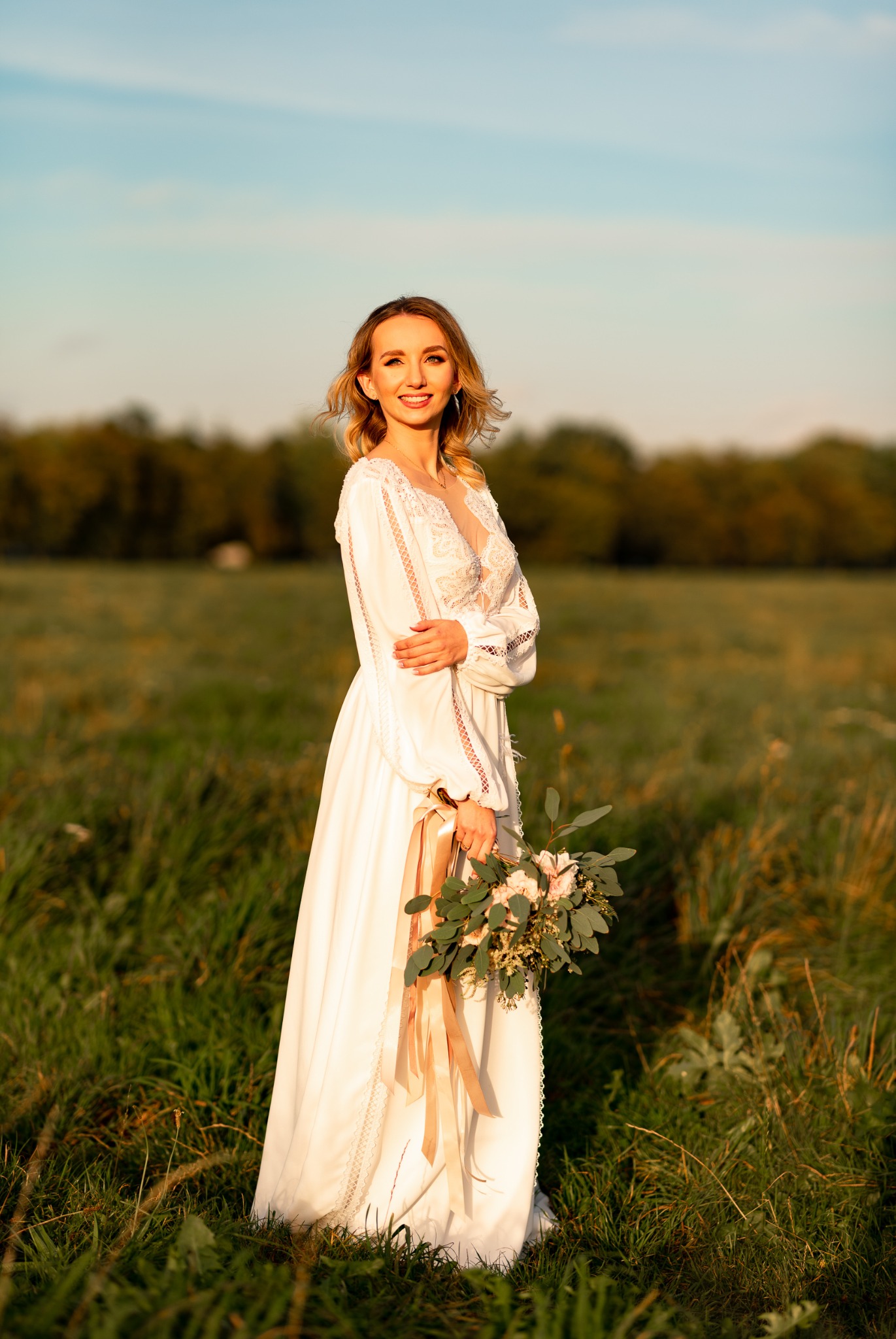 Panna młoda w białej sukni z koronkowymi detalami trzyma bukiet z różowymi kwiatami i zielonymi liśćmi, pozuje na łące o zachodzie słońca.