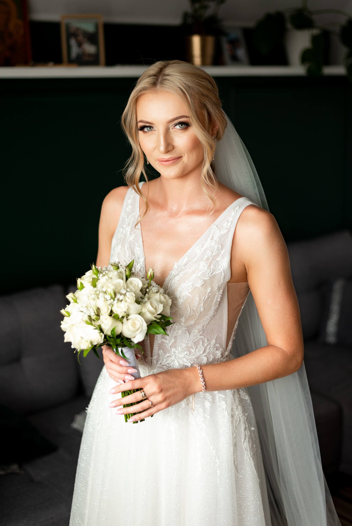 Panna młoda w białej sukni z koronkowymi detalami, trzymająca bukiet białych róż, z delikatnym makijażem i upiętymi blond włosami, pozująca na tle zielonej ściany i szarej sofy.