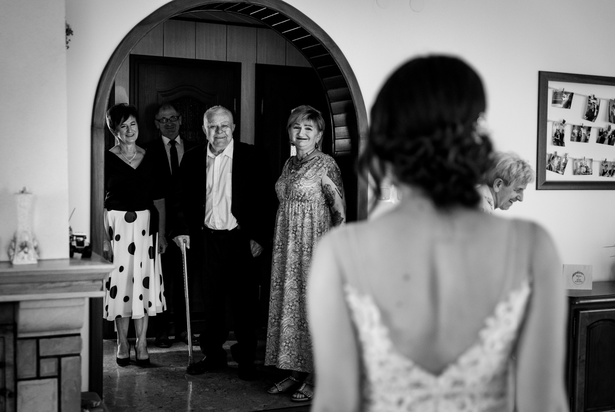 Panna młoda widziana od tyłu, przygotowująca się do ślubu, z rodziną w tle w drzwiach łukowych, czarno-biała fotografia.