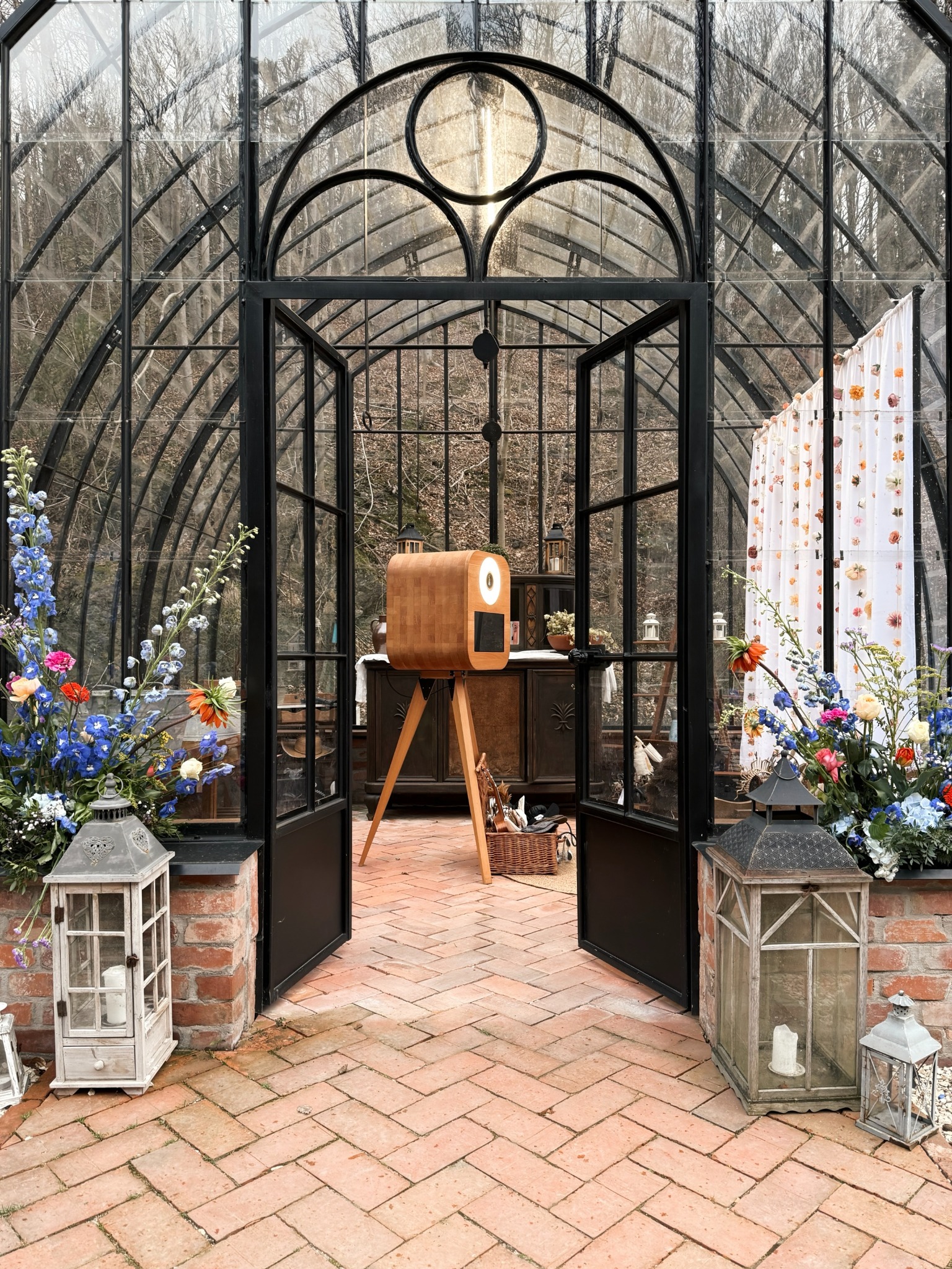 Drewniana fotobudka na trójnogu w szklarni z kwiatami i lampionami, Wrocław. Otwarta przestrzeń z ceglaną podłogą i dekoracjami, idealna na imprezy.