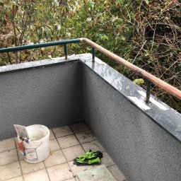 Dawid Ordon - Balkon po nałożeniu szarego tynku strukturalnego na ścianach, z widocznym wiadrem z zaprawą, kielnią i zielonymi rękawicami roboczymi na podłodze z płytek. Zabezpieczone taśmą malarską obrzeża...