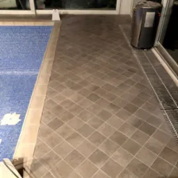 Wykończona podłoga z płytek ceramicznych w odcieniach szarości, ułożonych w karo, przylegająca do basenu z niebieską mozaiką. Widoczny grzejnik obok okna z aluminiową kratką odpływową.