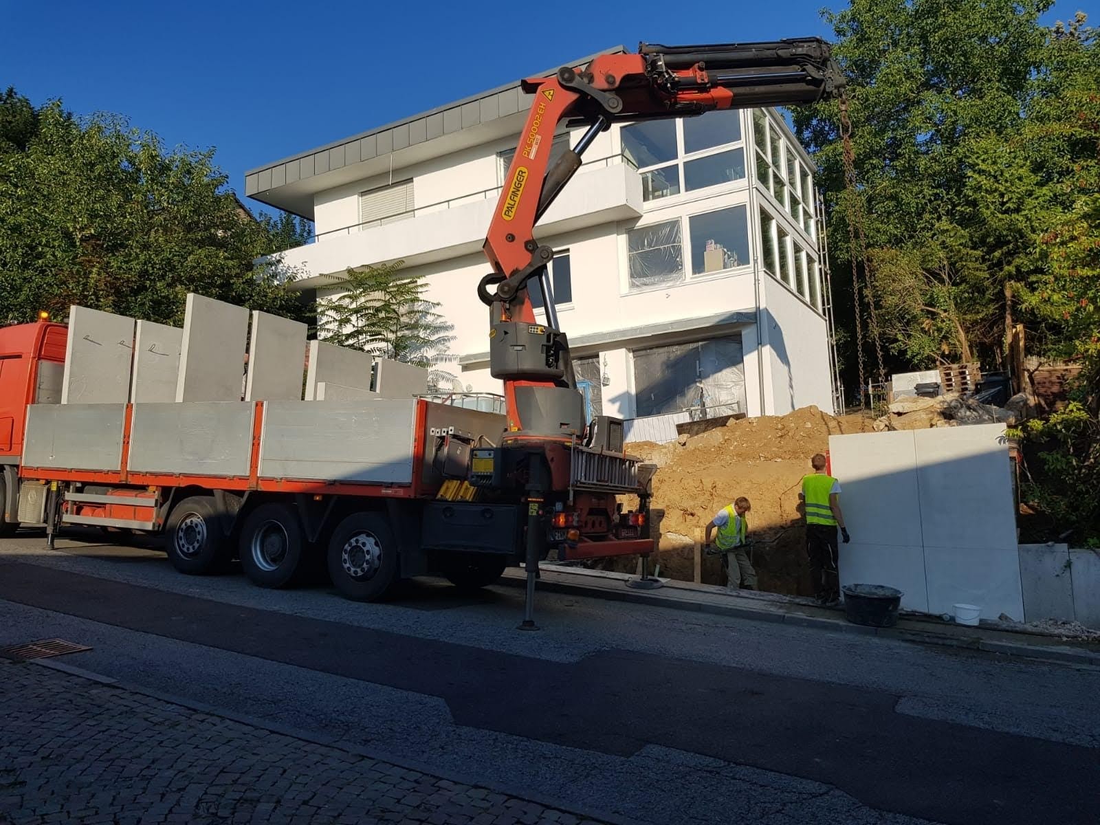 Montaż prefabrykowanych elementów betonowych przy użyciu dźwigu Palfinger na budowie domu jednorodzinnego, dwóch pracowników w kamizelkach odblaskowych nadzoruje proces.