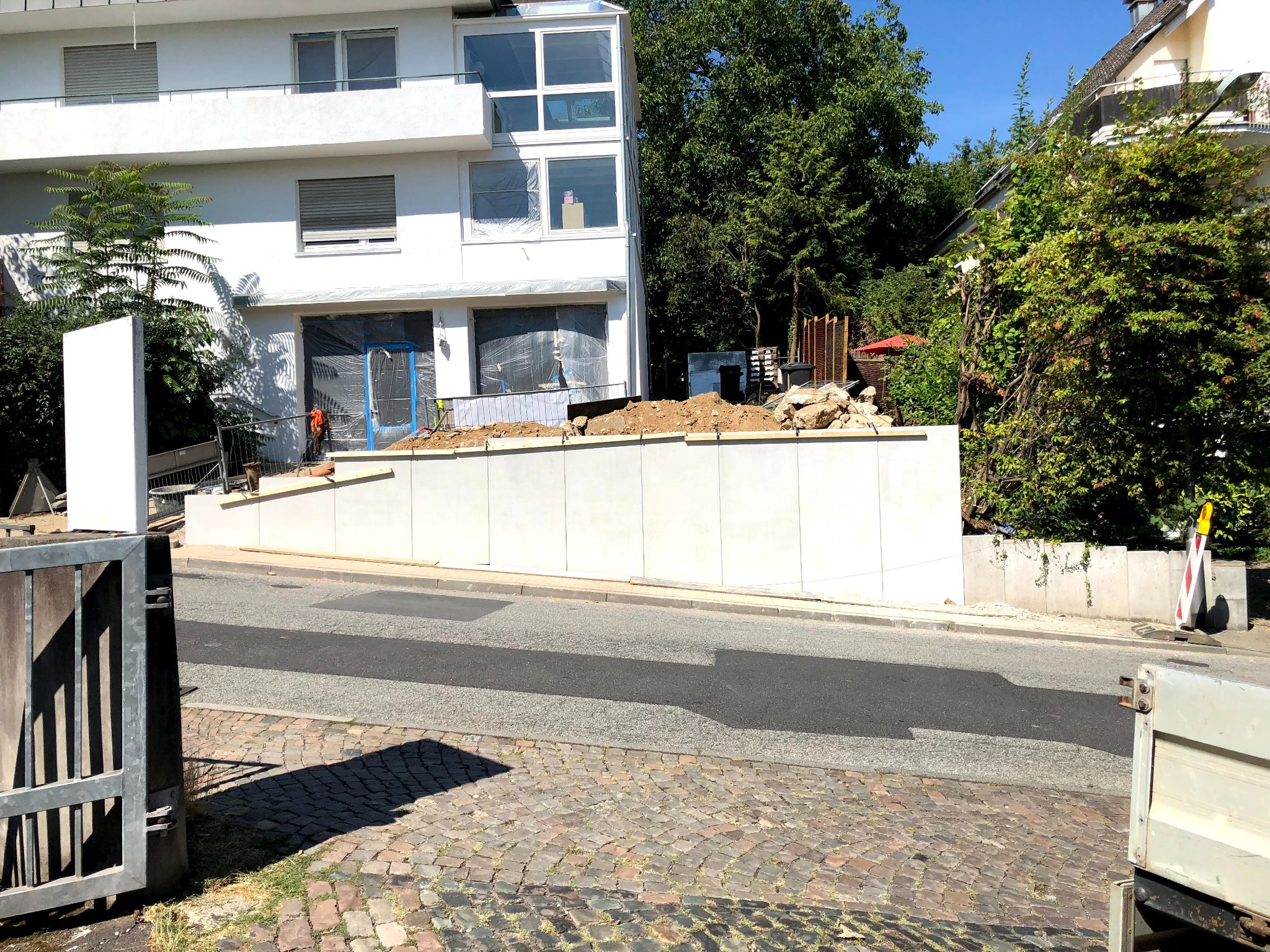 Biały, nowoczesny dom w trakcie remontu, z zabezpieczonymi oknami folią, widoczne materiały budowlane na podwyższeniu i fragment ulicy z kostki brukowej.