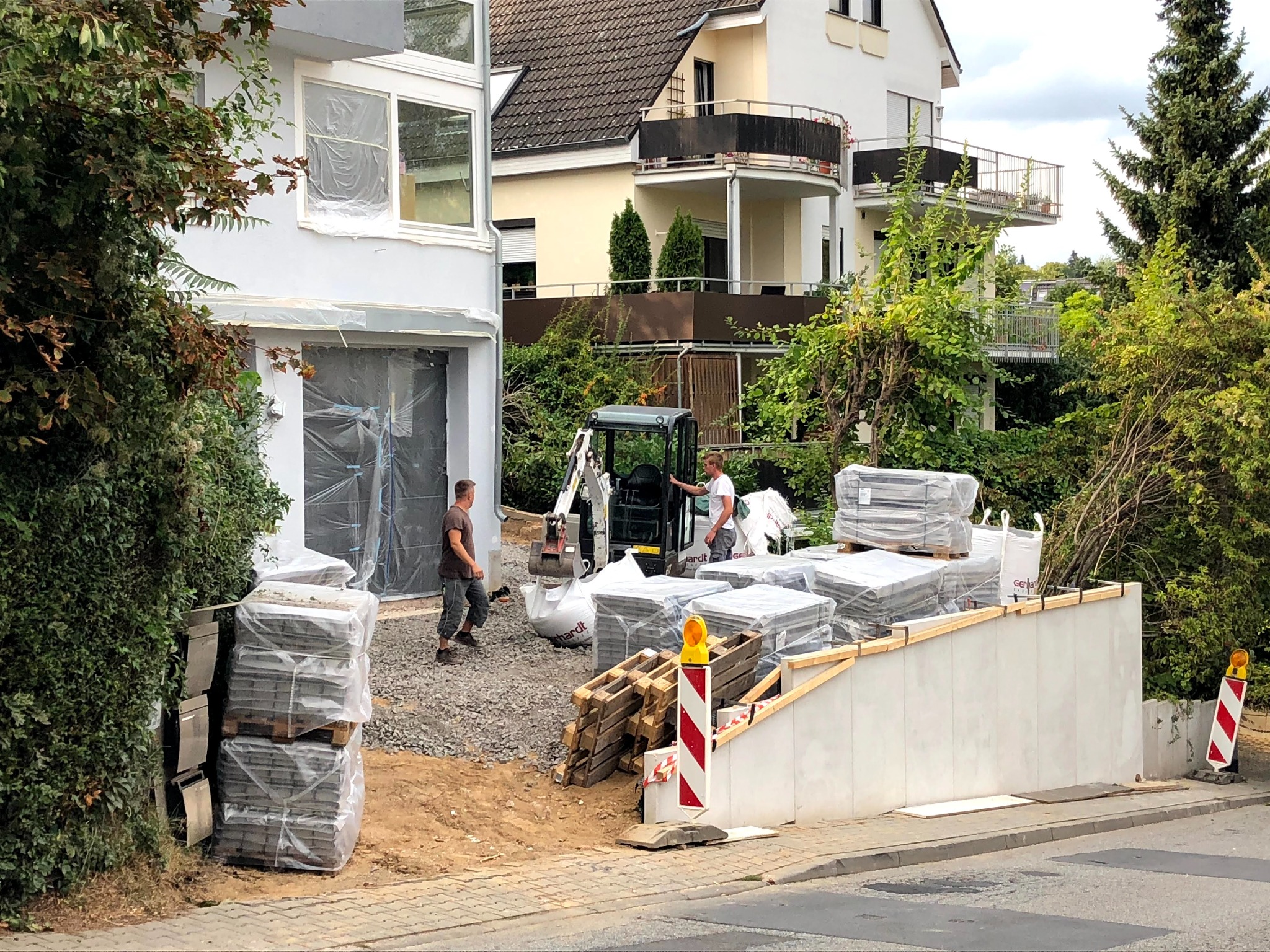 Dwaj mężczyźni pracują przy układaniu kostki brukowej przed budynkiem mieszkalnym, używając małej koparki. Okna budynku zabezpieczone folią malarską. Stosy kostki brukowej na paletach i luzem.