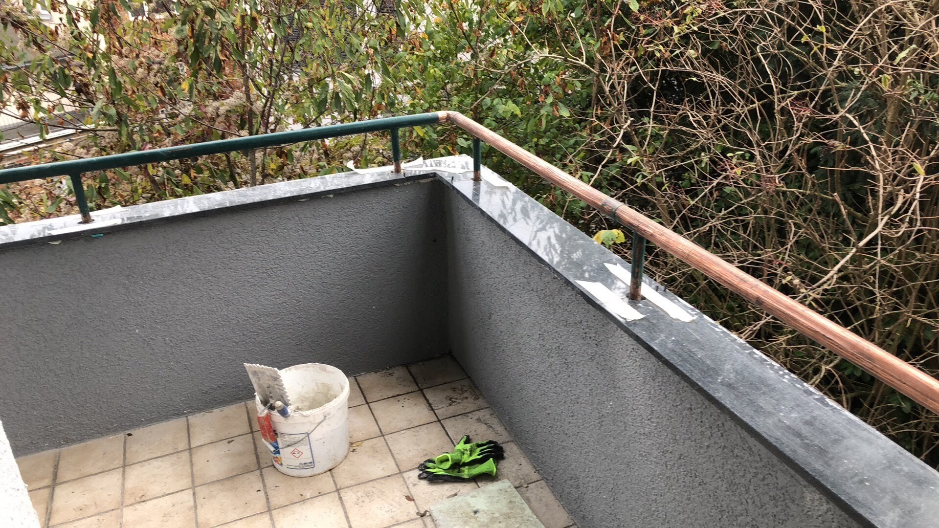 Balkon po nałożeniu szarego tynku strukturalnego na ścianach, z widocznym wiadrem z zaprawą, kielnią i zielonymi rękawicami roboczymi na podłodze z płytek. Zabezpieczone taśmą malarską obrzeża...