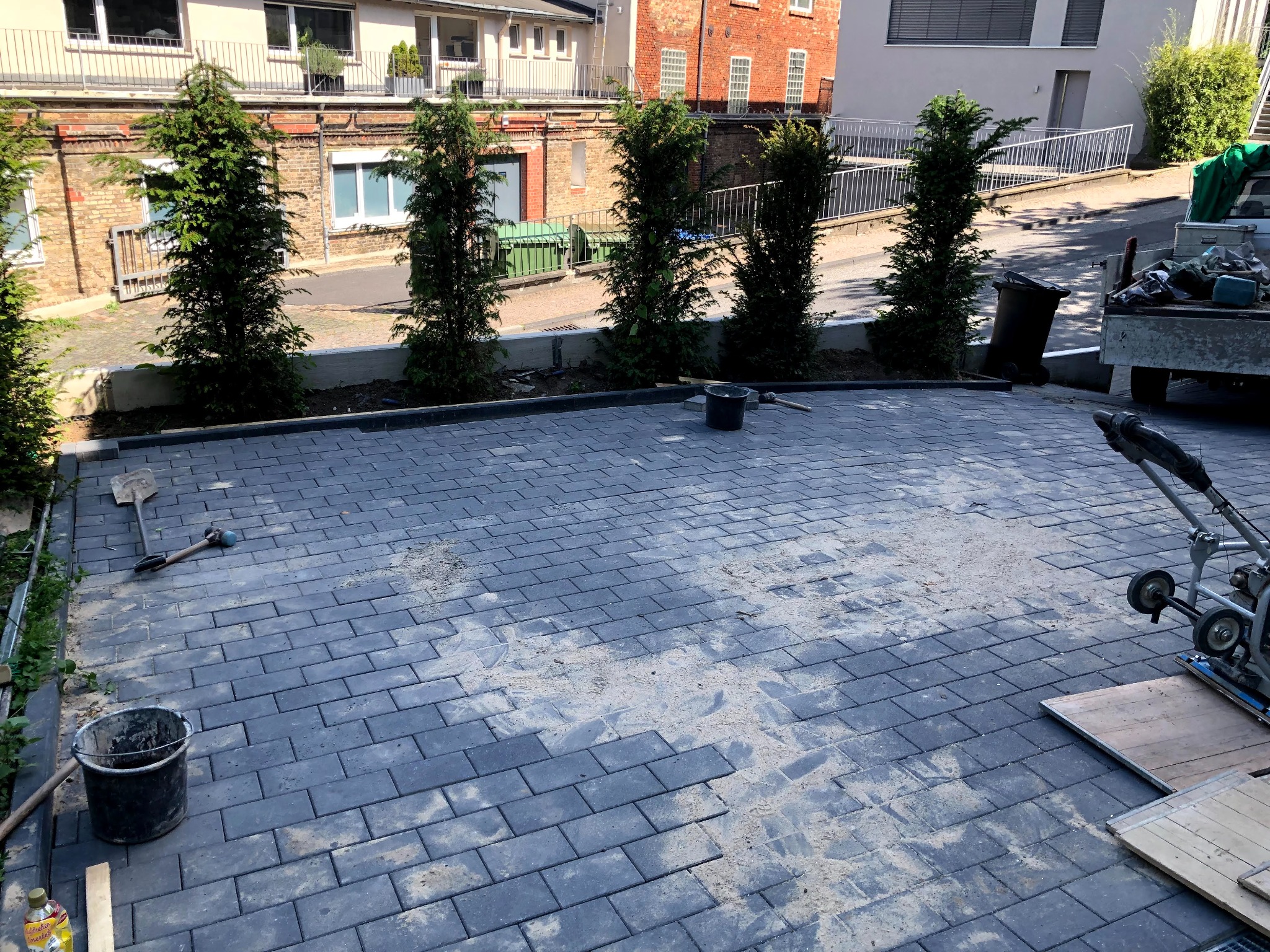 Układanie szarej kostki brukowej na podjeździe, widoczna ubijarka wibracyjna, wiadro, łopata i młotek, w tle tuje i budynki.