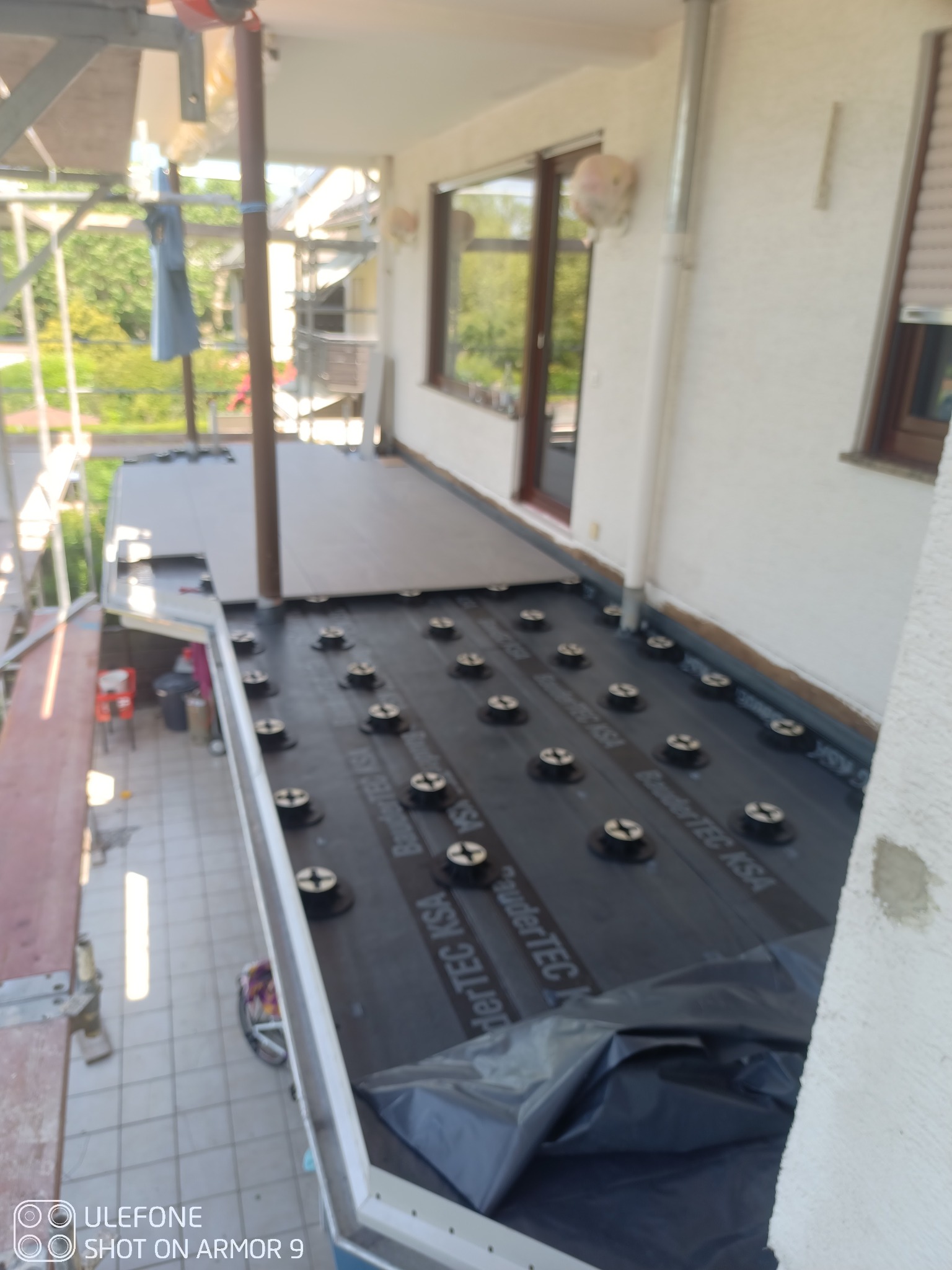 Montaż izolacji przeciwwodnej na balkonie, widoczne membrany z mocowaniami, częściowo ułożone płyty podłogowe, rusztowanie w tle.