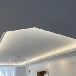 Nowoczesny sufit podwieszany z oświetleniem LED w kształcie trapezu, widoczny fragment żyrandola z kulistymi kloszami i punktowe oświetlenie w białym pomieszczeniu.