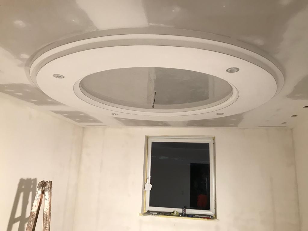 Okrągły sufit podwieszany z oświetleniem punktowym w trakcie montażu, widoczne ślady szpachlowania na suficie i ścianach, drabina aluminiowa oparta o ścianę przy oknie.
