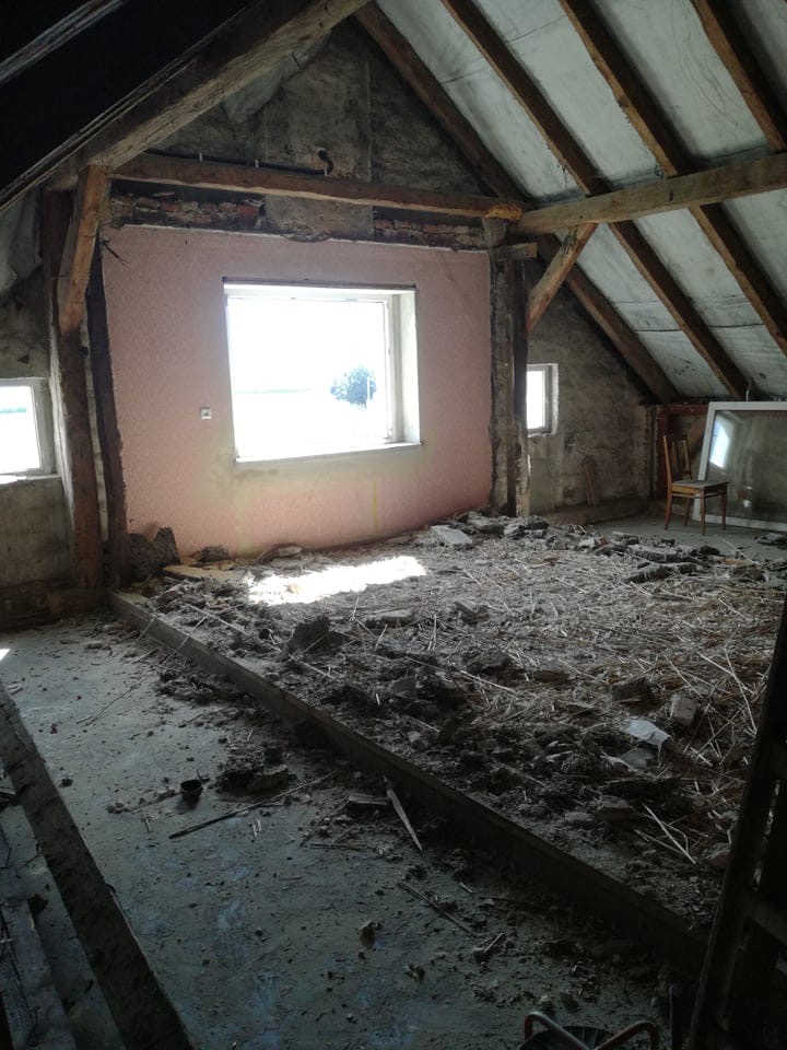 Poddasze w trakcie remontu: ściana z oknem częściowo pokryta różową gładzią, widoczne drewniane belki konstrukcyjne, gruz i resztki materiałów na podłodze, w tle krzesło i rama okienna.