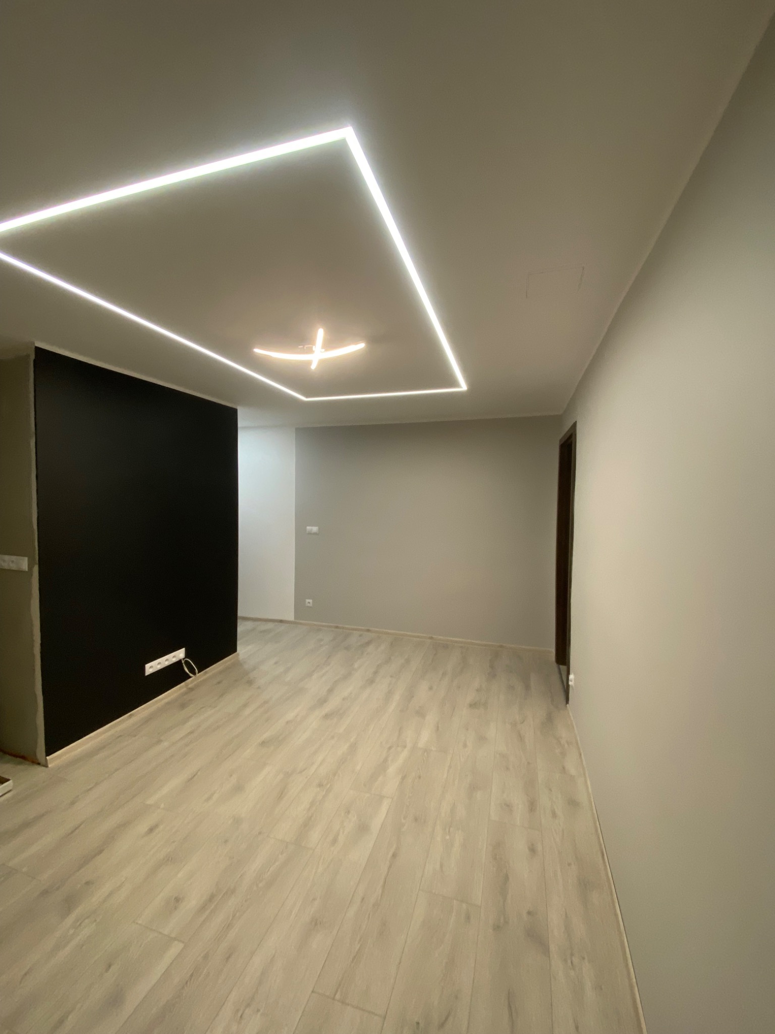 Wnętrze pokoju z nowoczesnym oświetleniem sufitowym w kształcie kwadratu i krzyża, czarną ścianą z gniazdkami i panelami podłogowymi o jasnym odcieniu drewna.