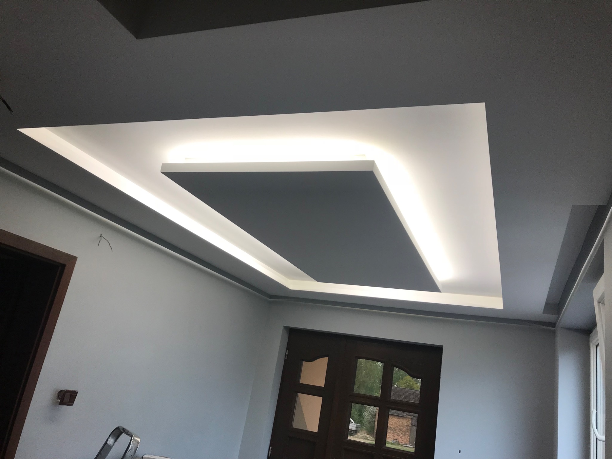 Nowoczesny sufit podwieszany z oświetleniem LED, szare ściany, widoczne drzwi z szybami i fragment drabiny malarskiej.