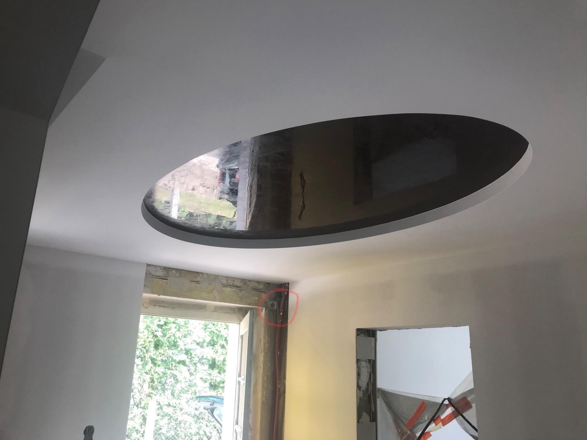 Okragły otwór w suficie podwieszanym podczas remontu, widoczne przygotowane ściany i okno z widokiem na zieleń.