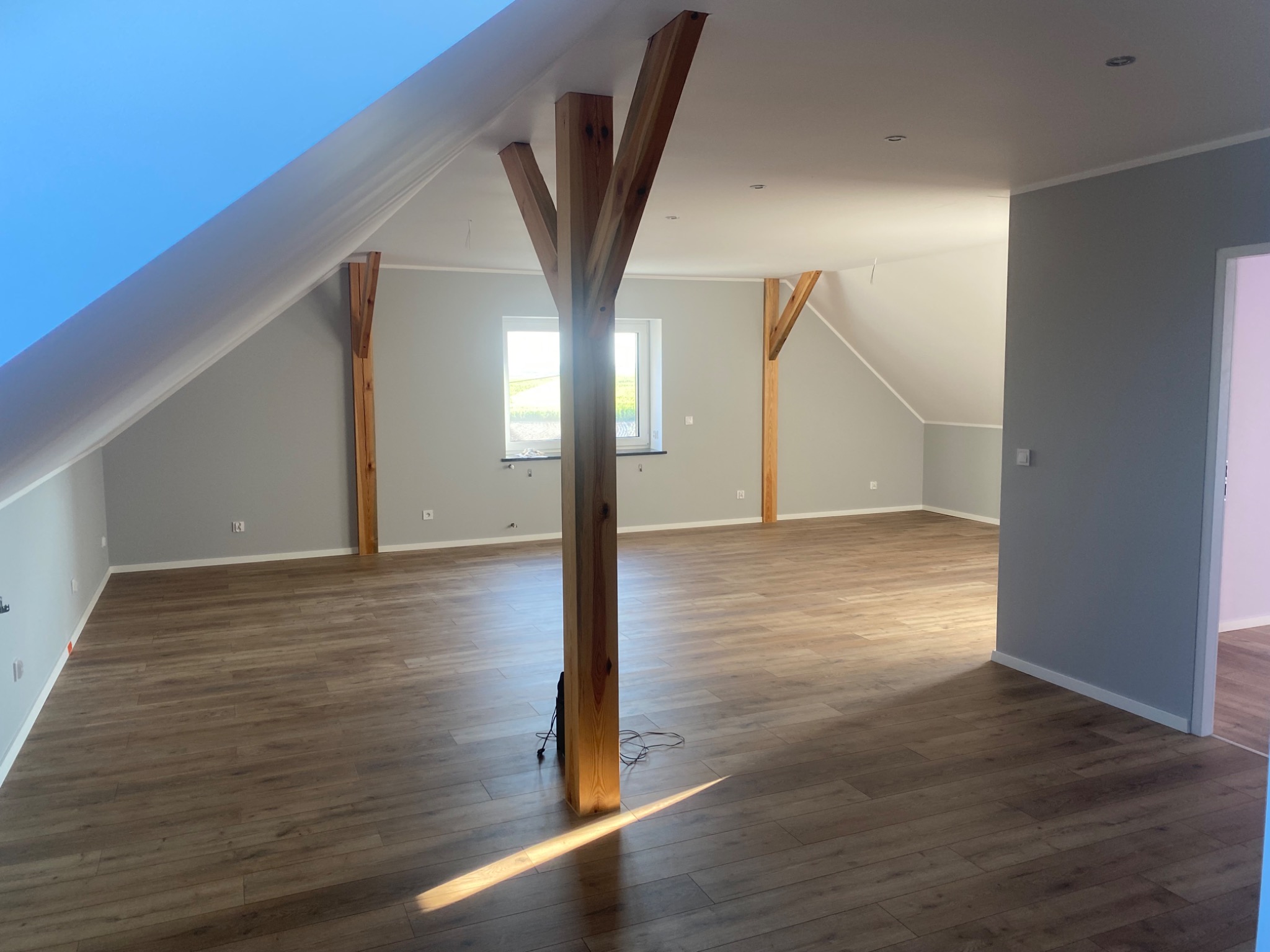 Przestronne, jasne poddasze po remoncie z drewnianymi belkami konstrukcyjnymi, panelami podłogowymi w odcieniach brązu i szarymi ścianami. Widoczne okno z widokiem na zewnątrz i punktowe...