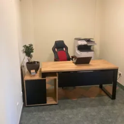 Nowoczesne biuro z drewnianym biurkiem o metalowych nogach, czarnym frontem, gamingowym krzesłem, laptopem i drukarką w tle, uzupełnione małą roślinką doniczkową.