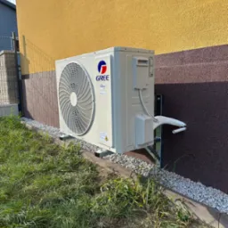 Zewnętrzna jednostka klimatyzacji marki Gree zamontowana na elewacji budynku z dwukolorową fasadą (żółty tynk i brązowy kamień dekoracyjny) i widocznymi przewodami w osłonie. Urządzenie posadowione...