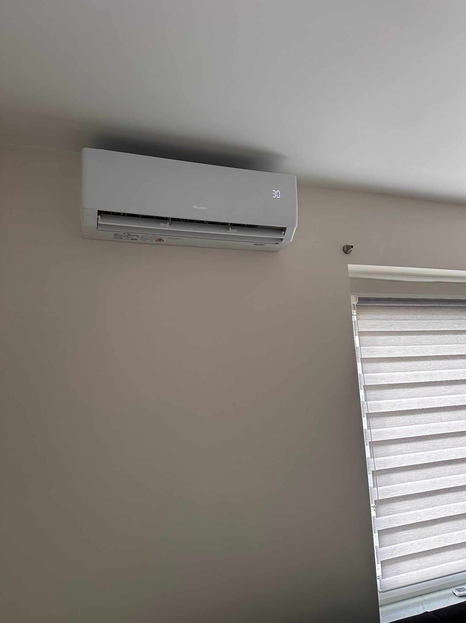 Biały klimatyzator marki Gree zamontowany na ścianie w pomieszczeniu w Pszczynie, obok okna z żaluzjami poziomymi; na wyświetlaczu widoczna temperatura 30 stopni.