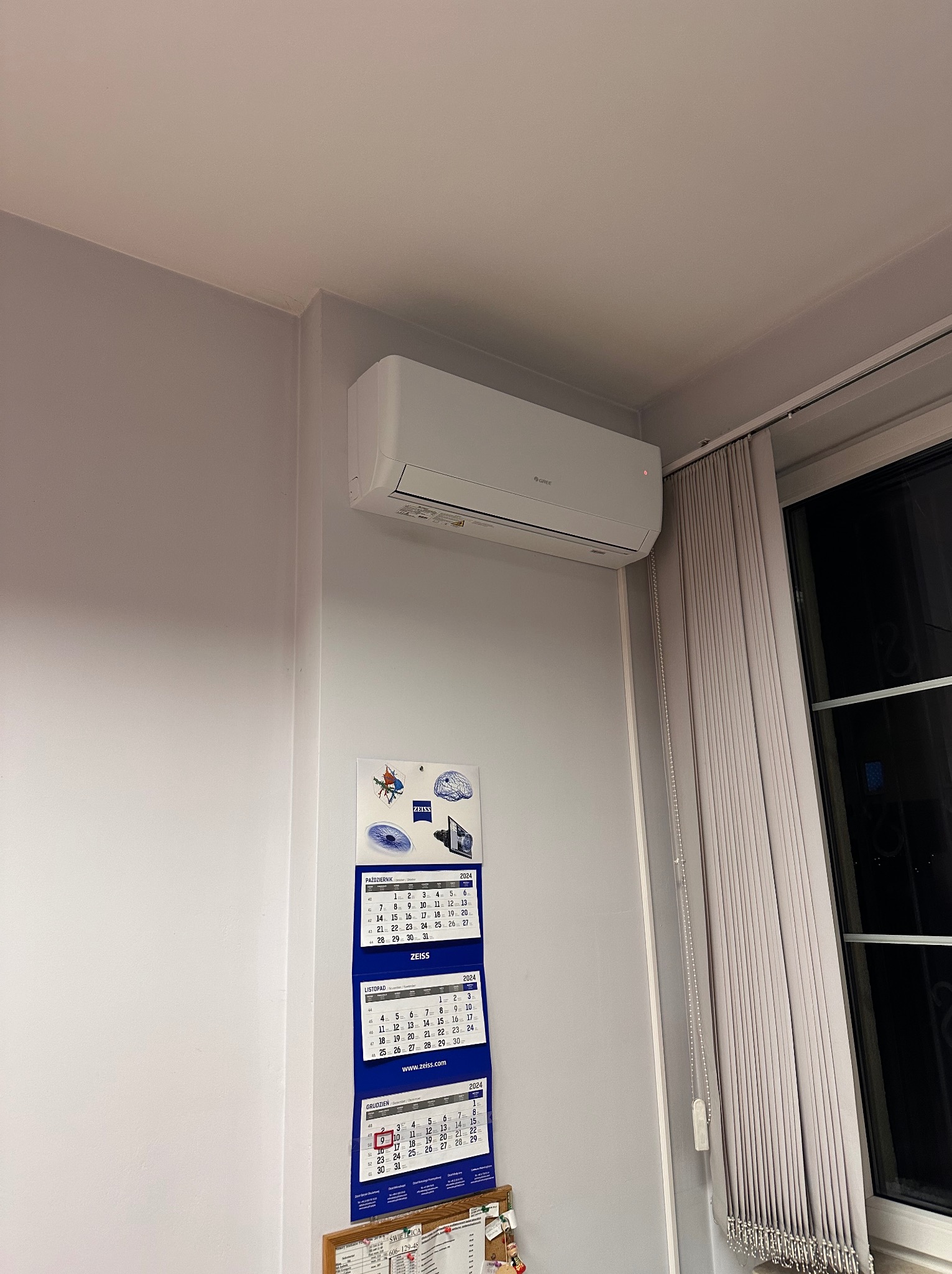Biały klimatyzator ścienny marki Gree zamontowany w pomieszczeniu z kalendarzem Zeiss na ścianie obok okna z zasłoniętymi żaluzjami.