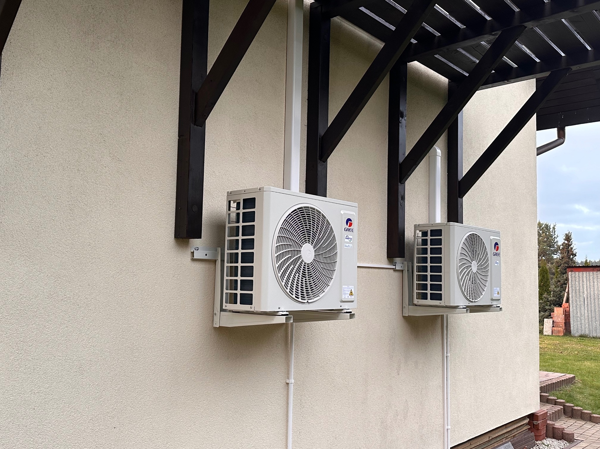 Dwa identyczne klimatyzatory marki Gree zamontowane na jasnej elewacji domu, widoczne wsporniki i rury odprowadzające skropliny, zadaszenie z drewnianych belek nad urządzeniami.