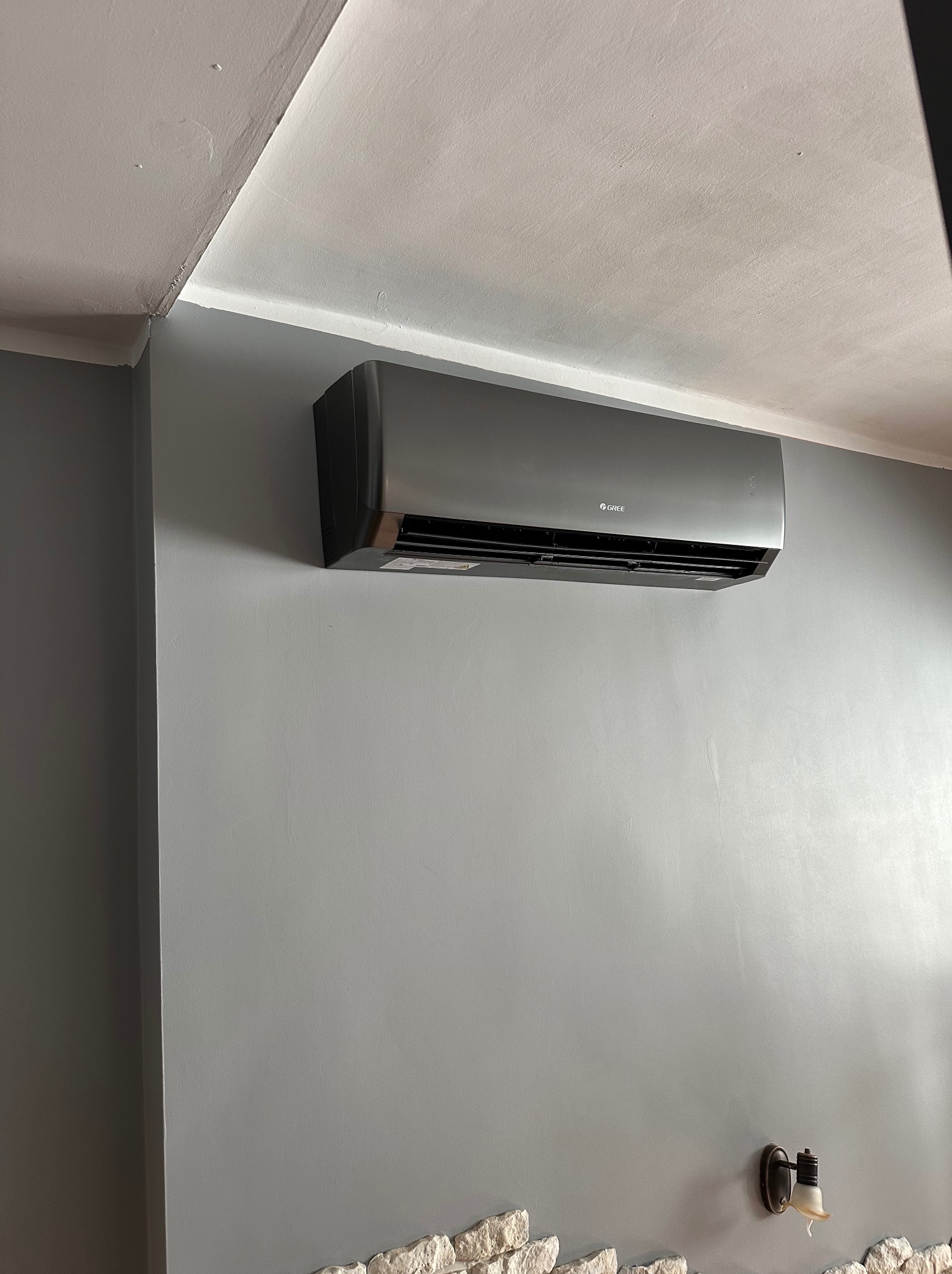 Szary klimatyzator marki Gree zamontowany na szarej ścianie w pomieszczeniu z dekoracyjnym panelem imitującym kamień u dołu i lampką ścienną.