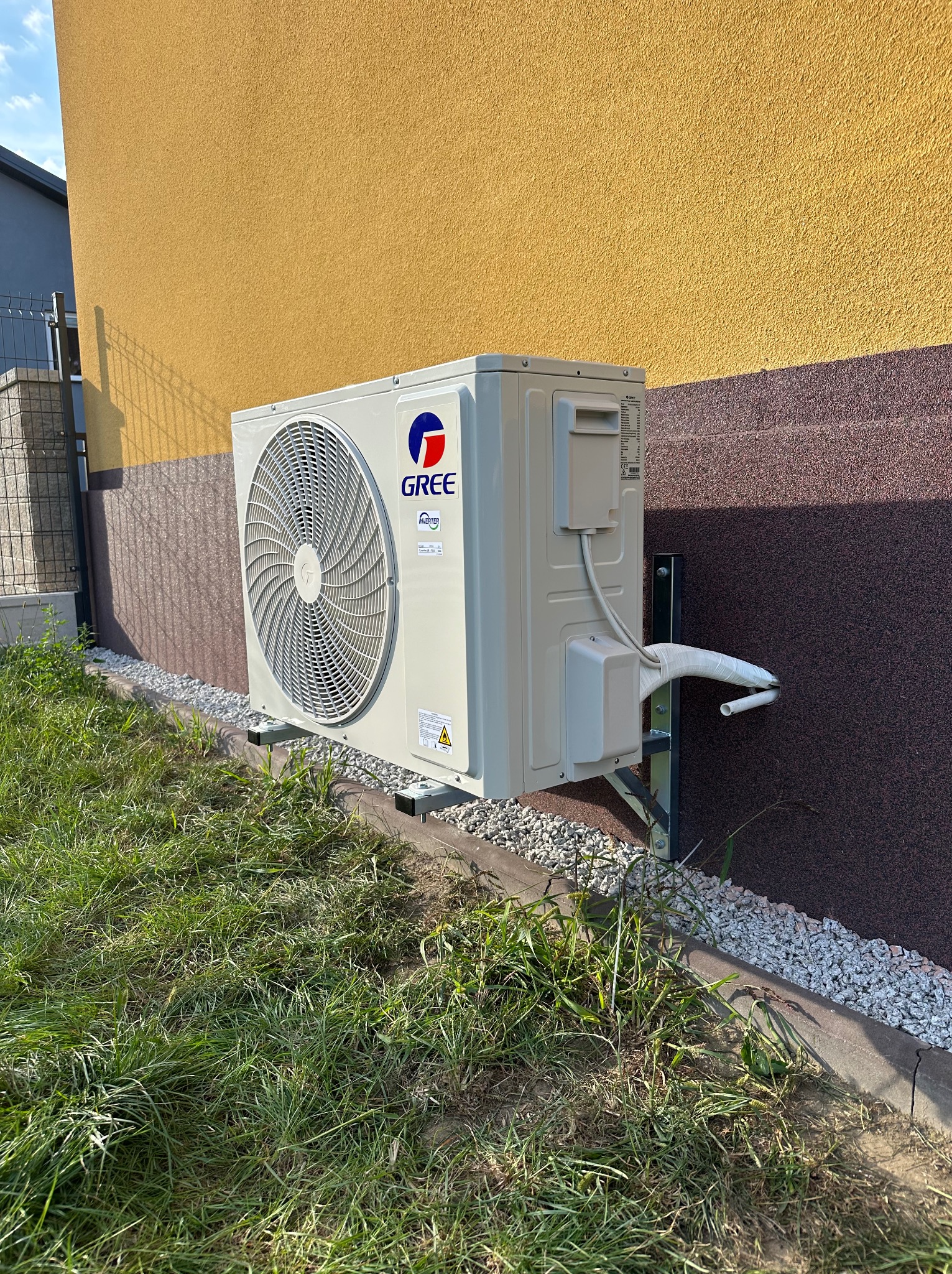 Zewnętrzna jednostka klimatyzacji marki Gree zamontowana na elewacji budynku z dwukolorową fasadą (żółty tynk i brązowy kamień dekoracyjny) i widocznymi przewodami w osłonie. Urządzenie posadowione...